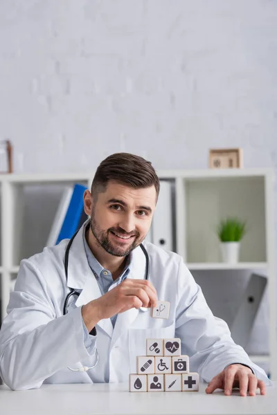 Medico felice guardando la fotocamera mentre fa piramide con simboli medici sulla scrivania — Foto stock