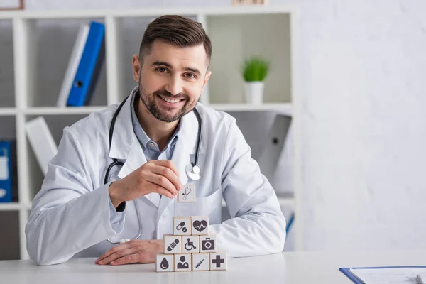 Médecin en manteau blanc souriant près de la pyramide des cubes avec des icônes médicales sur le bureau — Photo de stock