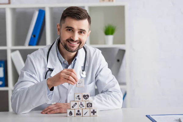 Physician in white coat smiling near pyramid of cubes with medical icons on desk — Stock Photo