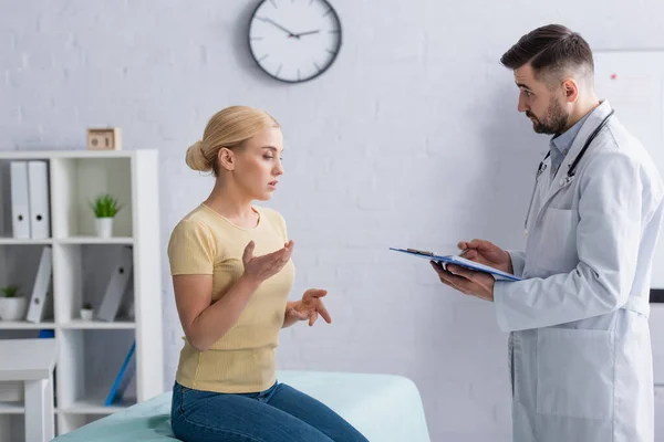 Woman sitting on medical couch and talking to doctor with clipboard — Stock Photo