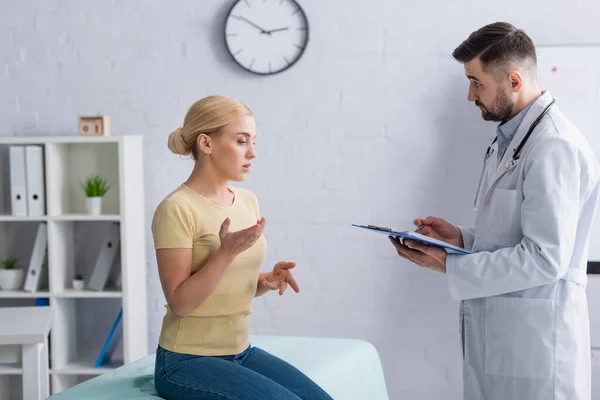 Mujer sentada en el sofá médico y hablando con el médico con el portapapeles - foto de stock