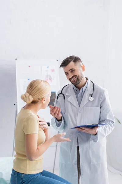 Lächelnder Arzt mit Klemmbrett im Gespräch mit blonder Frau in Klinik — Stockfoto