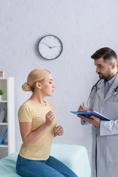 Médico escribiendo en portapapeles cerca de la mujer rubia sentada en el sofá médico - foto de stock