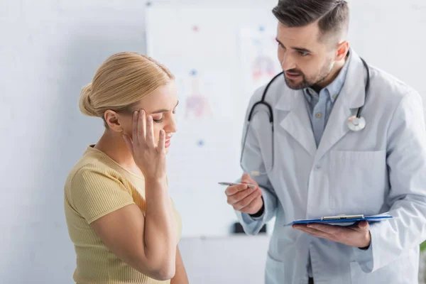 Doctor with clipboard talking to woman touching head with closed eyes — Stock Photo