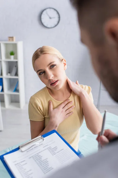 Frau berührt Hals und Brust nahe verschwommenem Arzt mit Klemmbrett — Stockfoto