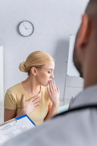 Mujer rubia tosiendo con los ojos cerrados cerca de médico borroso - foto de stock