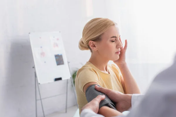 Mujer rubia que sufre de dolor de cabeza mientras que el médico pone el manguito de tonómetro en su brazo - foto de stock