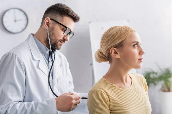 Arzt in weißem Mantel und Brille untersucht blonde Frau mit Stethoskop — Stockfoto