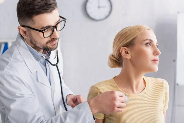 Doctor en anteojos examinando mujer rubia con estetoscopio - foto de stock