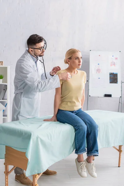 Femme en jeans assis sur le canapé médical tandis que le médecin l'examine avec stéthoscope — Photo de stock