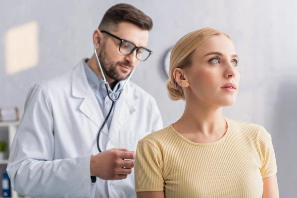 Doctor en anteojos examinando mujer rubia con estetoscopio - foto de stock