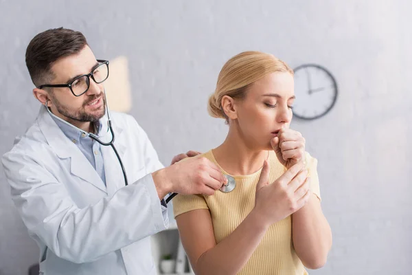 Mujer enferma tosiendo cerca del médico examinándola con estetoscopio en la clínica - foto de stock