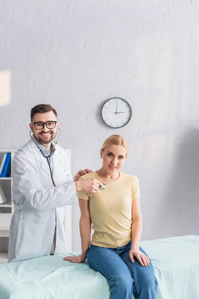 Arzt mit Brille blickt in Kamera, während er blonde Frau mit Stethoskop untersucht — Stockfoto