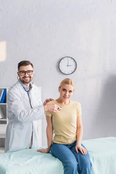 Médico en gafas de vista mirando a la cámara mientras examina mujer rubia con estetoscopio - foto de stock