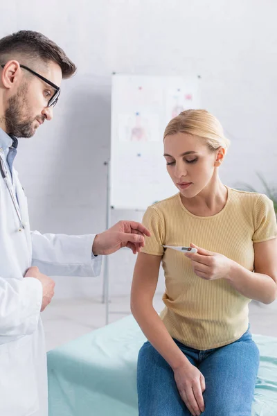 Doctor dando termómetro electrónico a la mujer sentada en el sofá médico - foto de stock