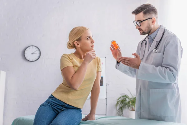 Médico apuntando a las píldoras mientras habla con la mujer rubia sentada en el sofá médico - foto de stock