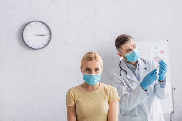 Mujer rubia con máscara de seguridad mirando la cámara cerca del médico llenando la jeringa con la vacuna - foto de stock