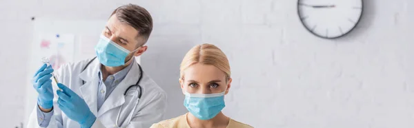 Frau in medizinischer Maske blickt in die Kamera, während Arzt Spritze mit Impfstoff füllt, Banner — Stockfoto