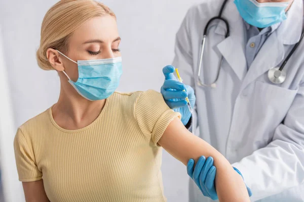 Médico en guantes de látex y bata blanca dando inyección de vacuna a la mujer en máscara protectora - foto de stock