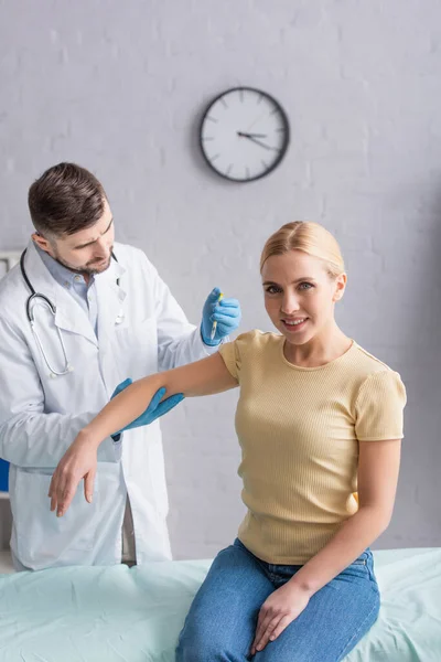 Médecin en manteau blanc et gants en latex vaccinant femme souriant à la caméra à la clinique — Photo de stock