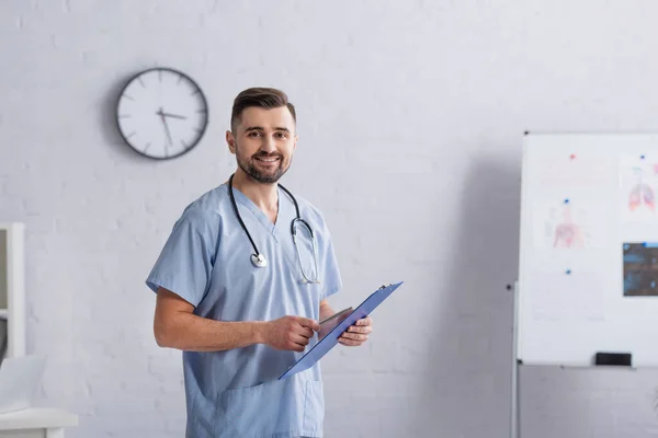 Medico sorridente in uniforme blu guardando la fotocamera mentre in piedi con appunti — Foto stock