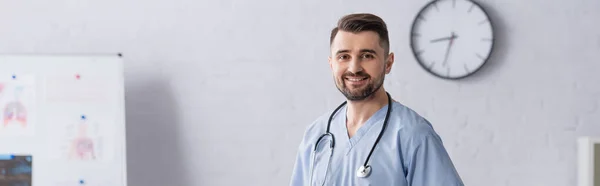 Médico feliz vestindo uniforme azul e estetoscópio no pescoço olhando para a câmera na clínica, bandeira — Fotografia de Stock