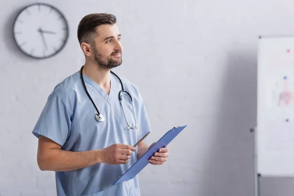 Lächelnder Arzt in blauer Uniform schaut weg, während er Klemmbrett hält — Stockfoto