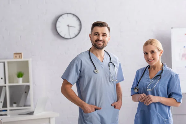 Médico de pé com as mãos em bolsos de uniforme perto colega sorridente — Fotografia de Stock