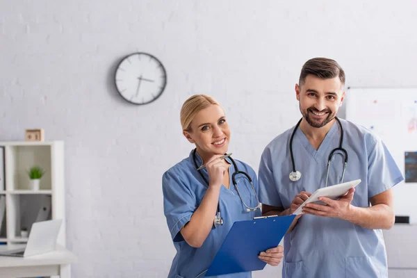Positive physicians with clipboard and digital tablet looking at camera in hospital — Stock Photo