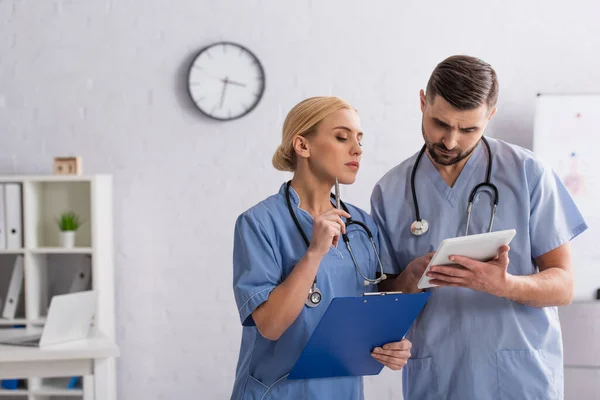 Médicos reflexivos en uniforme azul mirando tableta digital en el hospital - foto de stock