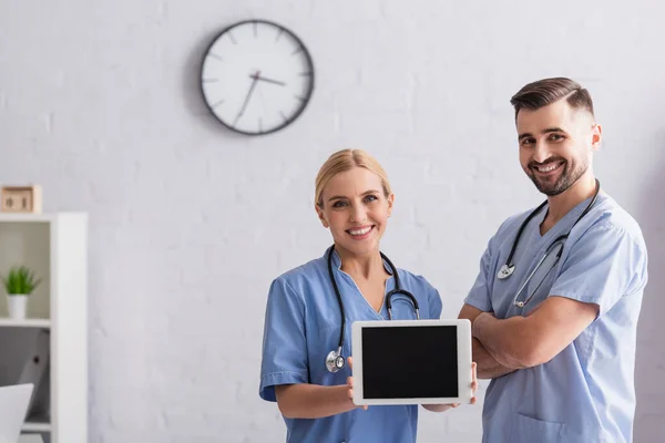 Весёлая медсестра показывает цифровую таблетку с чистым экраном рядом с положительным врачом, стоящим со скрещенными руками — стоковое фото