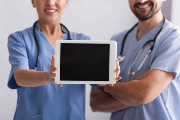 Vue recadrée d'un médecin souriant montrant une tablette numérique près d'un collègue aux bras croisés — Photo de stock