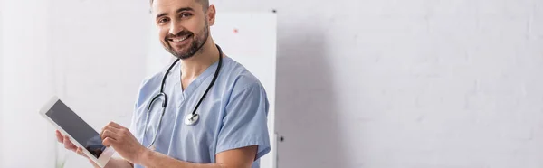 Радостный доктор в синей форме, держащий цифровой планшет с чистым экраном, баннер — стоковое фото