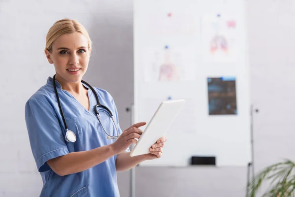 Médico rubia mirando a la cámara mientras usa la tableta digital en la clínica - foto de stock