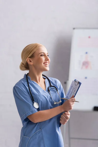 Cheerful doctor with clipboard and pen looking away in hospital — Stock Photo