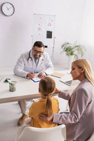 Pediatra sorrindo conversando com a menina com a mãe durante a consulta — Fotografia de Stock