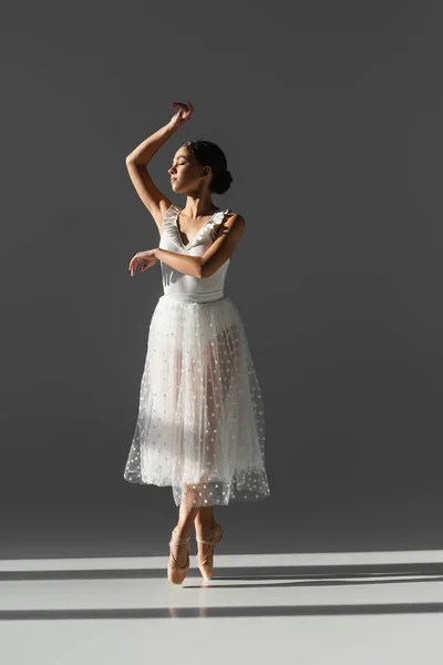 Vue latérale de la jeune ballerine aux yeux fermés dansant sur fond gris avec lumière du soleil — Photo de stock