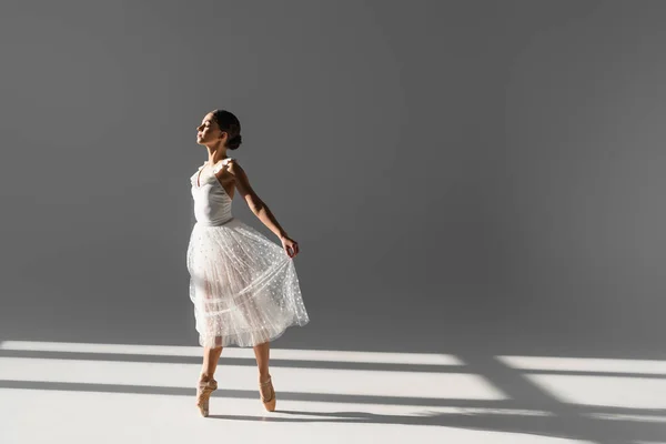 Vista lateral de la bailarina sosteniendo la falda mientras baila sobre fondo gris con luz solar - foto de stock