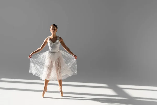 Full length of ballerina in skirt and pointe shoes dancing on grey background with sunlight — Stock Photo