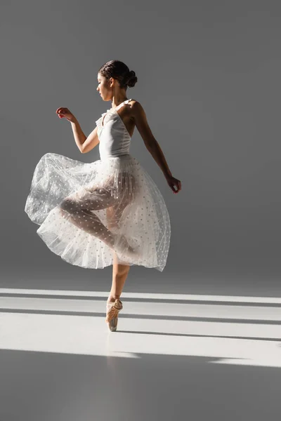 Seitenansicht einer eleganten Ballerina in weißem Rock, die auf grauem Hintergrund mit Sonnenlicht tanzt — Stockfoto