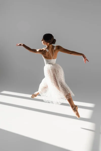 Side view of elegant ballerina jumping in sunlight on grey background — Stock Photo