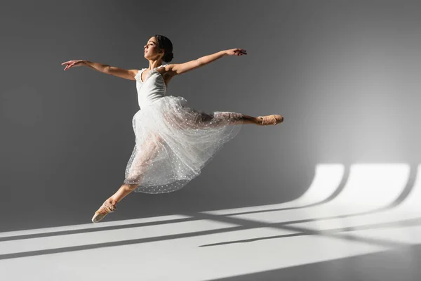 Vista lateral da bailarina em vestido branco pulando na luz solar no fundo cinza — Fotografia de Stock