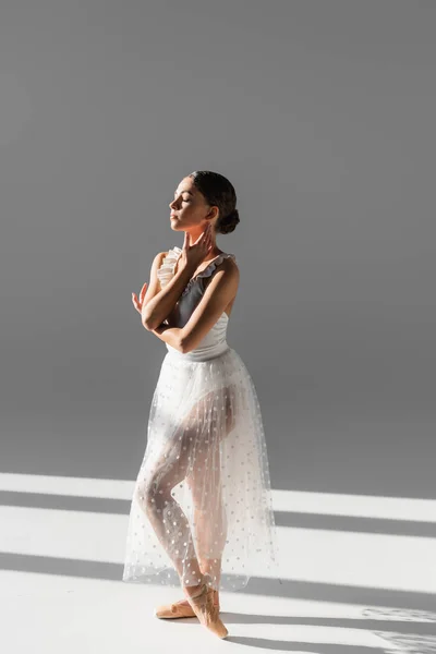 Side view of ballerina with closed eye touching neck on grey background with sunlight — Stock Photo