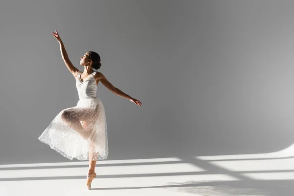 Side view of ballerina in white skirt standing on one leg on grey background with sunlight — Stock Photo