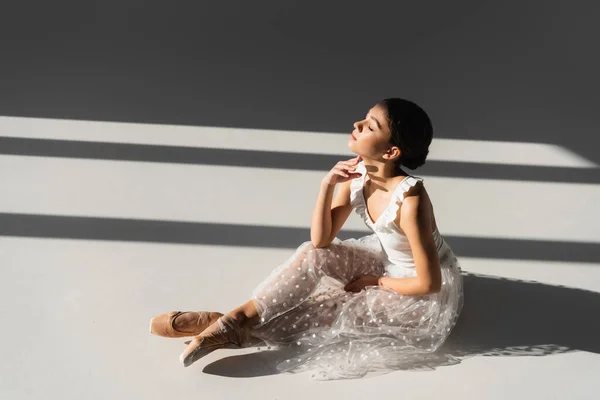 Vista lateral da bailarina com os olhos fechados sentados sobre fundo cinza com luz solar — Fotografia de Stock