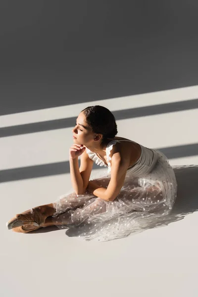 Side view of ballerina bending while sitting on grey background with sunlight — Stock Photo