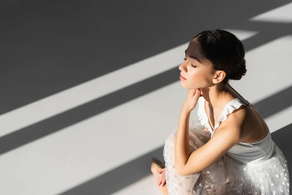 Professional ballerina sitting on grey background with sunlight — Stock Photo