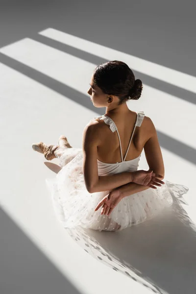 Side view of ballerina in white dress and pointe shoes sitting in sunlight on grey background — Stock Photo