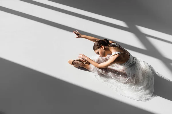 High angle view of pretty classical dancer bending on floor on grey background with sunlight — Stock Photo