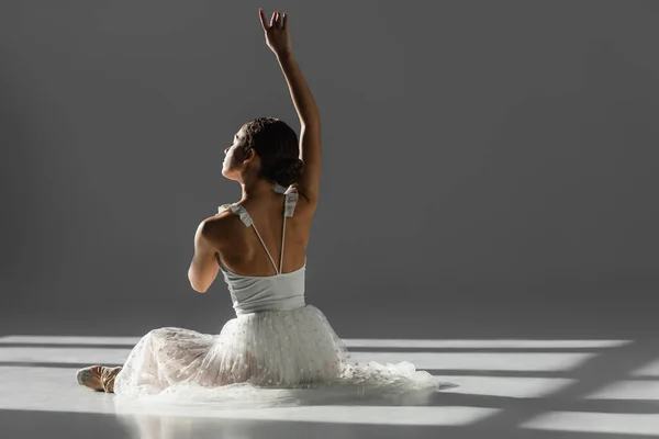 Ballerine levant la main tout en étant assis dans la lumière du soleil sur fond gris avec la lumière du soleil — Photo de stock